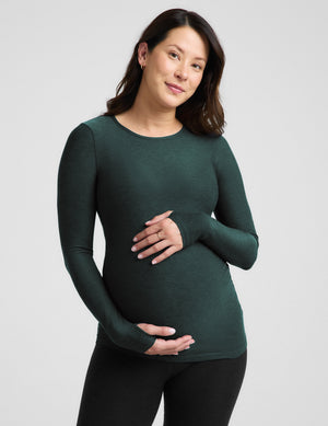 A pregnant woman wearing a dark forest green long-sleeve maternity top and black leggings, standing against a light gray studio background.