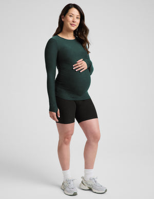 A pregnant woman modeling a dark green long-sleeve maternity top and black athletic shorts against a light gray studio background.