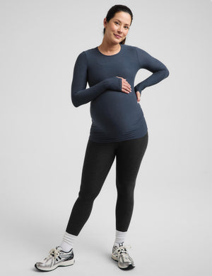 Pregnant woman wearing a blue maternity long sleeve top and black maternity leggings against a plain background. 