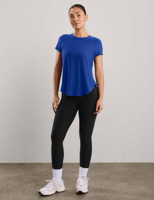 A woman modeling a short-sleeve athletic top in a solid royal blue color, paired with black leggings, against a neutral light gray studio background.