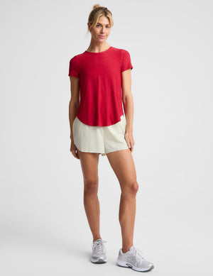 A woman wearing a bright red short-sleeve athletic top and light cream shorts against a light gray studio background.