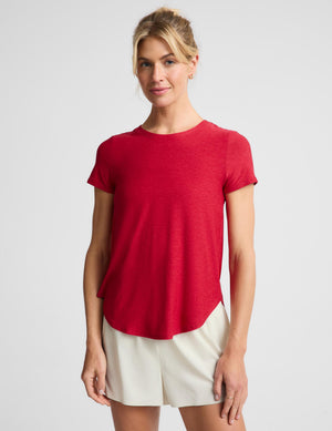 A woman modeling a short-sleeve crewneck t-shirt in heather red, paired with off-white shorts, against a light gray studio background.