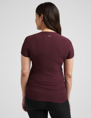 A pregnant woman wearing a short-sleeve crewneck maternity t-shirt in maroon and black bike shorts, standing against a light gray studio background.