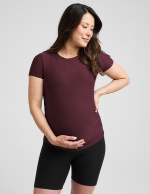 A pregnant woman wearing a short-sleeve crewneck maternity t-shirt in maroon and black bike shorts, standing against a light gray studio background.