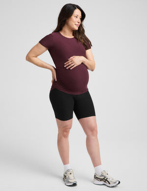 A pregnant woman wearing a short-sleeve crewneck maternity t-shirt in maroon and black bike shorts, standing against a light gray studio background.
