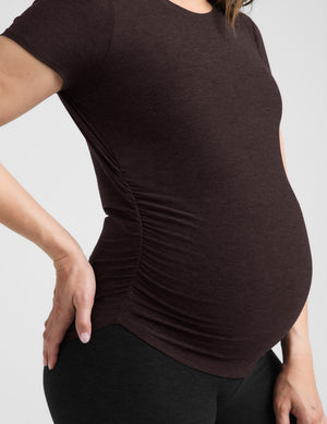 Pregnant woman wearing a brown maternity t-shirt and black leggings against a plain background. 