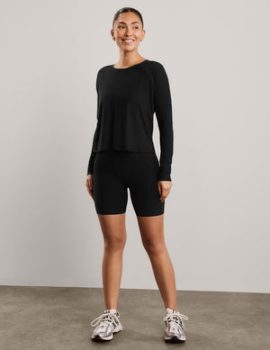A woman modeling a black long-sleeve crewneck top and black biker shorts against a plain light gray studio background.