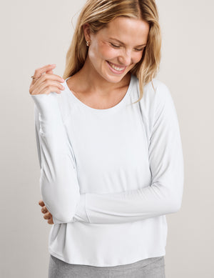 A woman modeling a white long-sleeve crewneck athletic top paired with heather gray leggings, set against a plain light gray studio background.