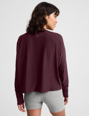 A woman wearing a dark burgundy long-sleeve crewneck top and heather gray bike shorts, standing against a light gray studio background.