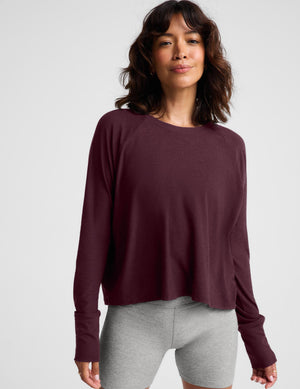 A woman wearing a dark burgundy long-sleeve crewneck top and heather gray bike shorts, standing against a light gray studio background.