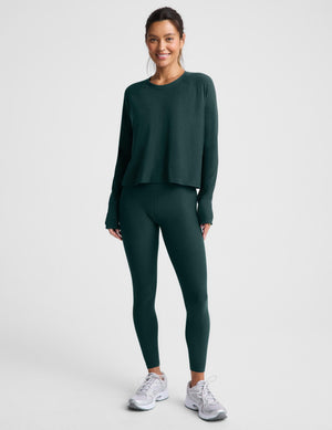 A woman modeling a dark forest green long-sleeve cropped top and matching leggings against a light gray studio background.