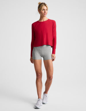 A woman wearing a bright red long sleeve and gray biker shorts against a plain background. 