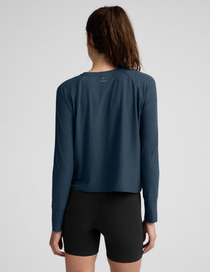 A woman modeling a dark slate blue long-sleeve cropped top with black bike shorts against a light gray studio background.