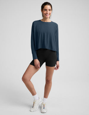 A woman modeling a dark slate blue long-sleeve cropped top with black bike shorts against a light gray studio background.