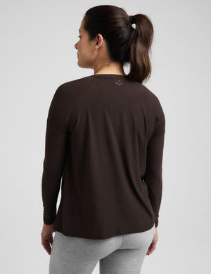 Pregnant woman wearing a brown long sleeve maternity top and gray leggings against a plain background. 