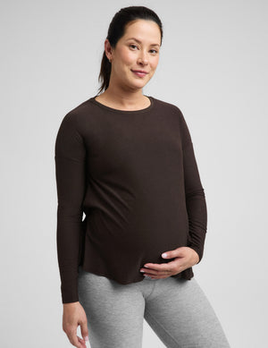 Pregnant woman wearing a brown long sleeve maternity top and gray leggings against a plain background. 