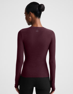 A woman modeling a long-sleeve crewneck top in a deep burgundy color, paired with black pants, against a solid light gray background.
