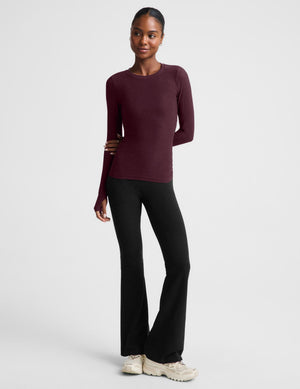 A woman modeling a long-sleeve crewneck top in a deep burgundy color, paired with black pants, against a solid light gray background.