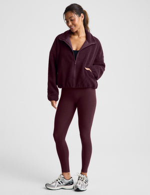 A woman wearing a dark burgundy boucle zip-up jacket and matching leggings, posing against a light gray studio background.