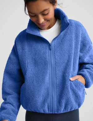 A woman wearing a textured fleece zip-up jacket in a medium blue color, paired with dark leggings, against a light gray studio background.