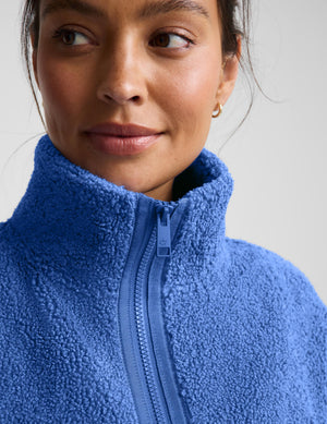 A woman wearing a textured fleece zip-up jacket in a medium blue color, paired with dark leggings, against a light gray studio background.