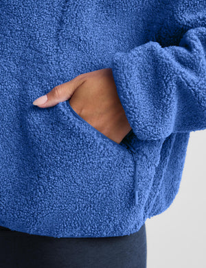 A woman wearing a textured fleece zip-up jacket in a medium blue color, paired with dark leggings, against a light gray studio background.