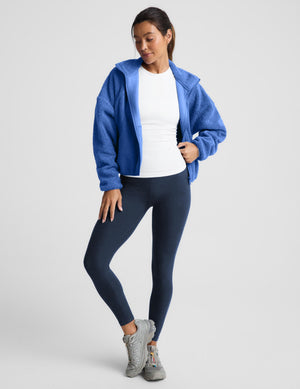 A woman wearing a textured fleece zip-up jacket in a medium blue color, paired with dark leggings, against a light gray studio background.