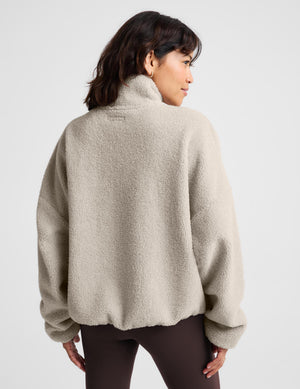 A woman modeling a light beige sherpa zip-up jacket over a dark brown sports bra and matching high-waisted leggings, paired with white sneakers, against a light gray studio background.