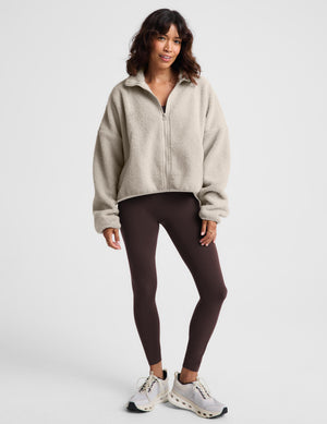 A woman modeling a light beige sherpa zip-up jacket over a dark brown sports bra and matching high-waisted leggings, paired with white sneakers, against a light gray studio background.