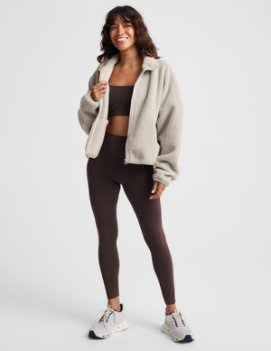 A woman modeling a light beige sherpa zip-up jacket over a dark brown sports bra and matching high-waisted leggings, paired with white sneakers, against a light gray studio background.