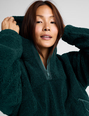 A woman wearing a dark green fleece half-zip pullover sweater and light gray leggings, standing against a light gray studio background.