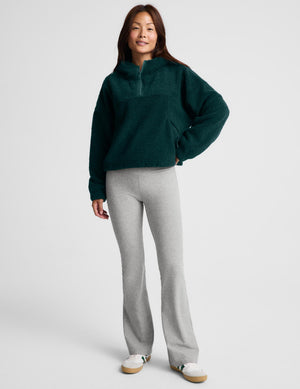 A woman wearing a dark green fleece half-zip pullover sweater and light gray leggings, standing against a light gray studio background.