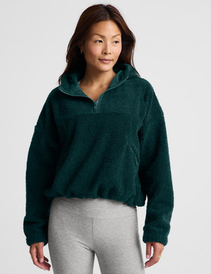A woman wearing a dark green fleece half-zip pullover sweater and light gray leggings, standing against a light gray studio background.