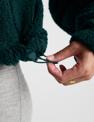 A woman wearing a dark green fleece half-zip pullover sweater and light gray leggings, standing against a light gray studio background.