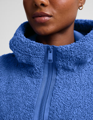 A woman wearing a textured blue fleece quarter-zip pullover sweater and gray leggings, posing against a light gray studio background.
