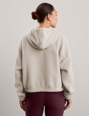 A woman modeling a light beige sherpa fleece half-zip pullover sweatshirt with a high collar, paired with dark burgundy leggings, against a solid light gray studio background.