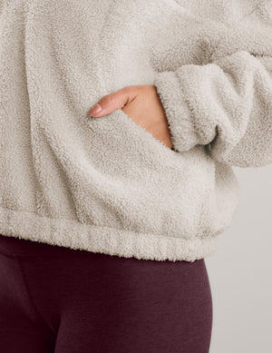 A woman modeling a light beige sherpa fleece half-zip pullover sweatshirt with a high collar, paired with dark burgundy leggings, against a solid light gray studio background.