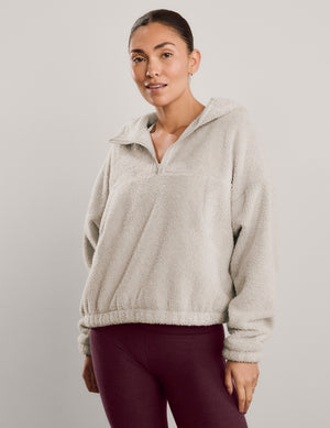 A woman modeling a light beige sherpa fleece half-zip pullover sweatshirt with a high collar, paired with dark burgundy leggings, against a solid light gray studio background.