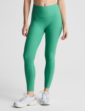 A woman modeling high-waisted green leggings and a white sports bra against a light gray studio background.