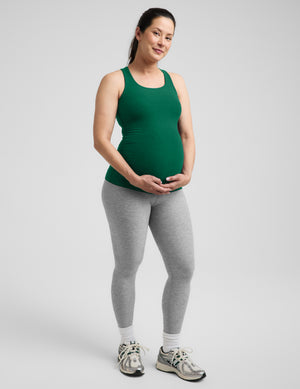 Pregnant woman wearing a green maternity racerback tank and gray leggings against a plain background. 