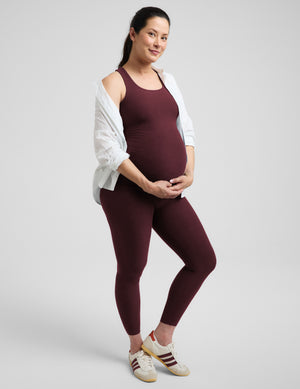 A pregnant woman posing in a matching burgundy activewear set consisting of a tank top and leggings, styled with an open white shirt and white sneakers, against a light gray studio background.