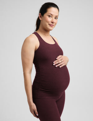 A pregnant woman wearing a matching dark burgundy maternity tank top and leggings set, standing against a solid light gray background.