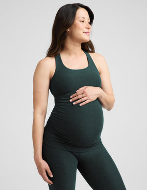 A pregnant woman wearing a dark forest green maternity tank top and matching leggings, standing against a light gray studio background.