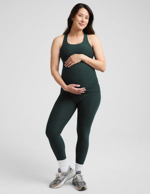 A pregnant woman wearing a dark forest green maternity tank top and matching leggings, standing against a light gray studio background.