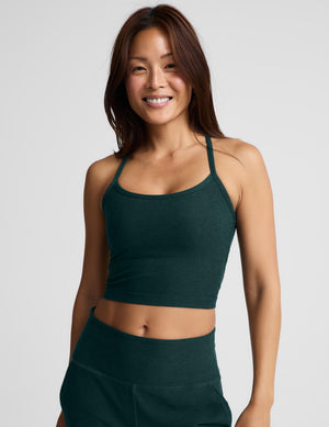 A smiling woman wearing a dark forest green athletic crop top and matching high-waisted pants against a light gray studio background.