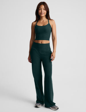 A smiling woman wearing a dark forest green athletic crop top and matching high-waisted pants against a light gray studio background.