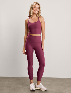 A smiling blonde woman wearing a matching heathered mauve activewear set, featuring a scoop-neck crop top and high-waisted leggings, against a solid light gray studio background.