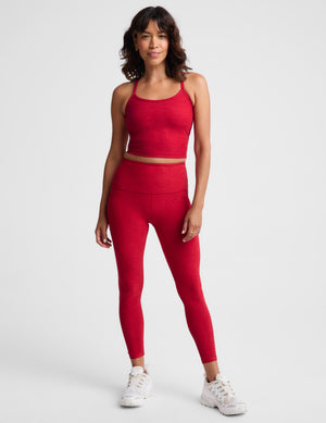 A woman wearing a matching red activewear set consisting of a cropped tank top and high-waisted leggings, photographed against a light gray studio background.