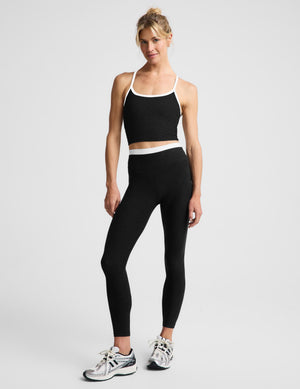 A woman modeling a black cropped tank and matching black leggings with white trim, photographed against a light gray studio background.