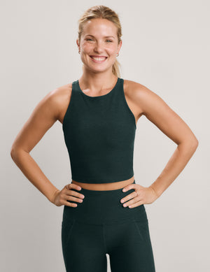 A smiling blonde woman posing in a dark forest green athletic crop top and matching high-waisted leggings against a light gray studio background.